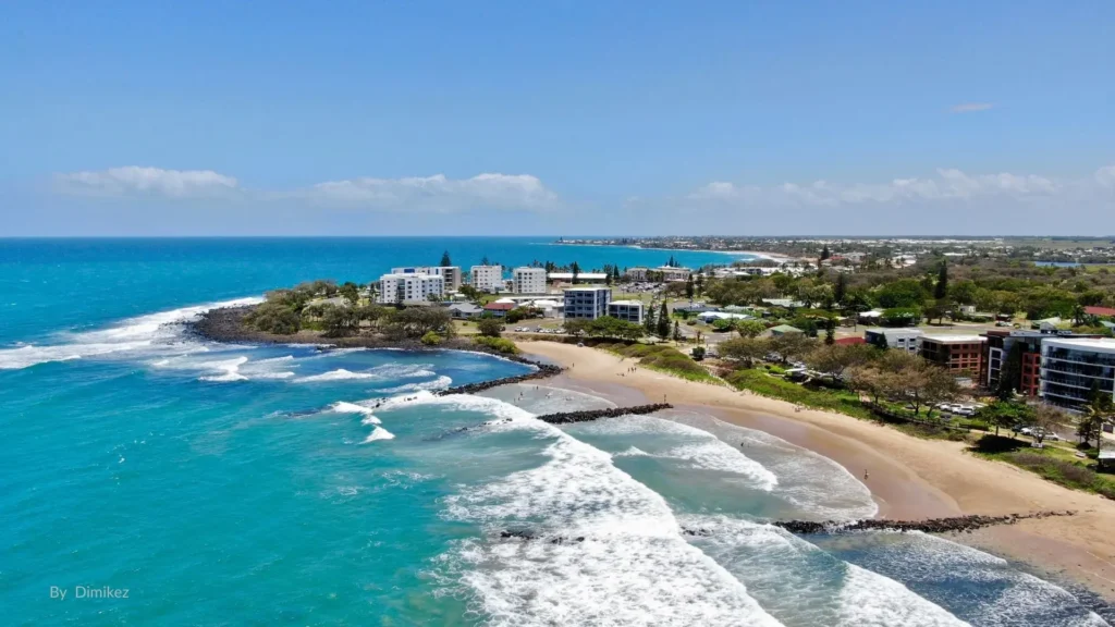 Aerial view of Bargara Beach, Bundaberg Queensland, showing sandy shoreline, surf waves, esplanade, and nearby resorts.