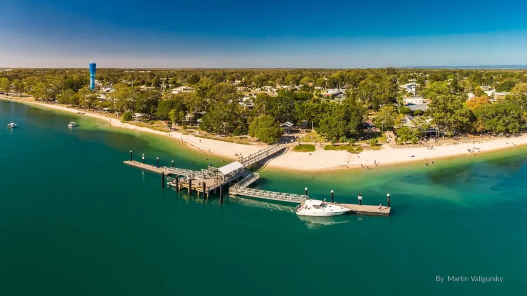 Aerial view of Bongaree Beach on Bribie Island, Queensland, showing the jetty, calm turquoise waters of Pumicestone Passage, sandy foreshore, and surrounding parklands
