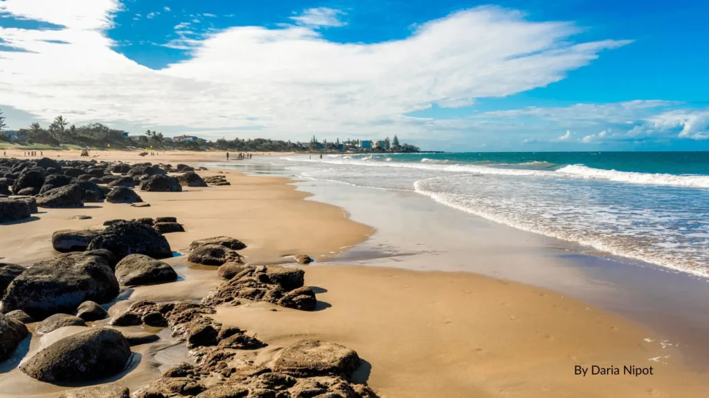 View of Kellys Beach, Bargara Queensland, showing sandy shoreline, calm ocean waves, rocky foreshore, and family-friendly beach setting