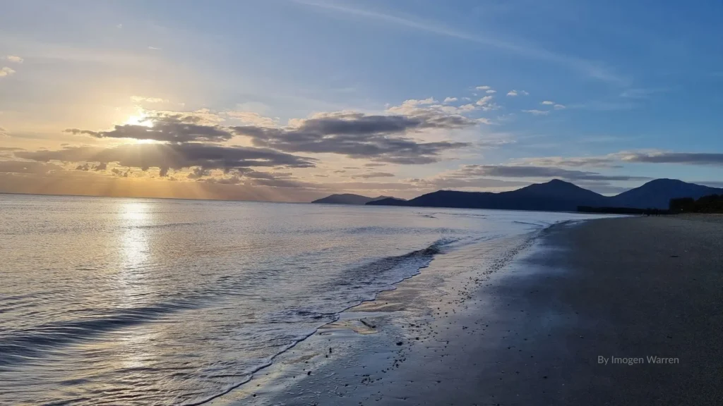 Sunset over Machans Beach in Cairns, Queensland, with calm waves, sandy shoreline, and mountain backdrop