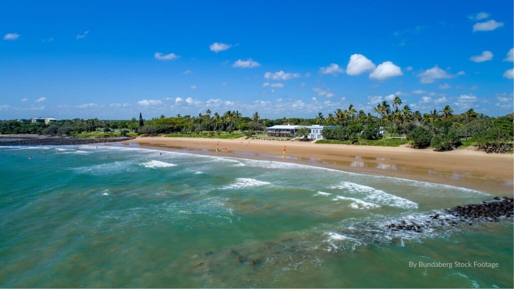 Nielson Park Beach, Bargara Queensland, showing golden sandy foreshore, grassy park areas, surf waves, and patrolled swimming zone