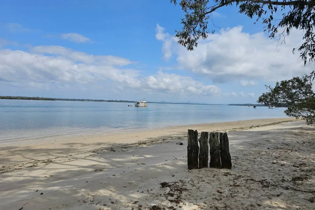 Peaceful shoreline at Banksia Beach, Bribie Island QLD, with calm Pumicestone Passage waters, anchored boats, and sandy foreshore framed by trees
