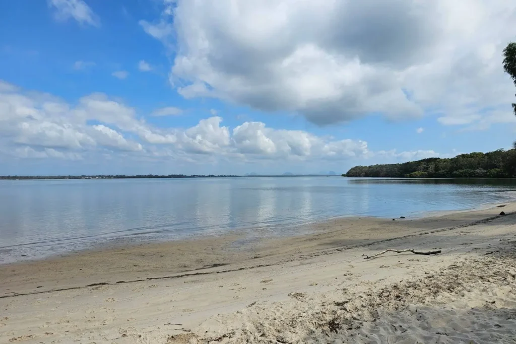 Wide sandy shoreline at Banksia Beach on Bribie Island, QLD, with calm blue waters of Pumicestone Passage and distant Glass House Mountains under a cloudy sky