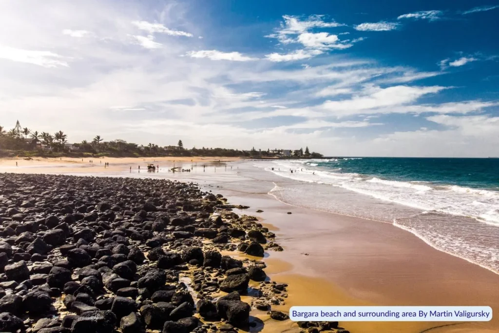 Scenic view of Bargara Beach in Queensland with black volcanic rocks in the foreground, golden sand stretching into the distance, gentle waves, and people walking under a partly cloudy sky.