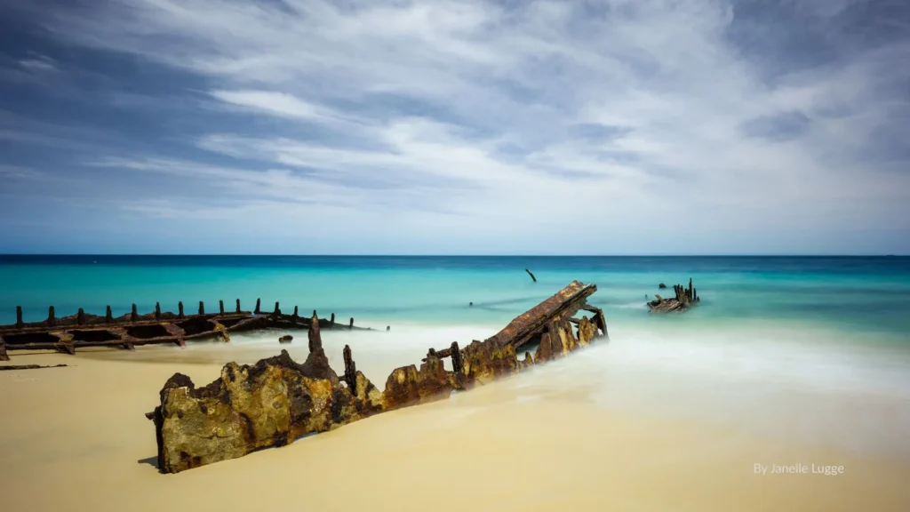 Rusting shipwreck remains on the sandy shoreline of Bulwer Beach with turquoise waters and cloudy skies, Moreton Island, Queensland.