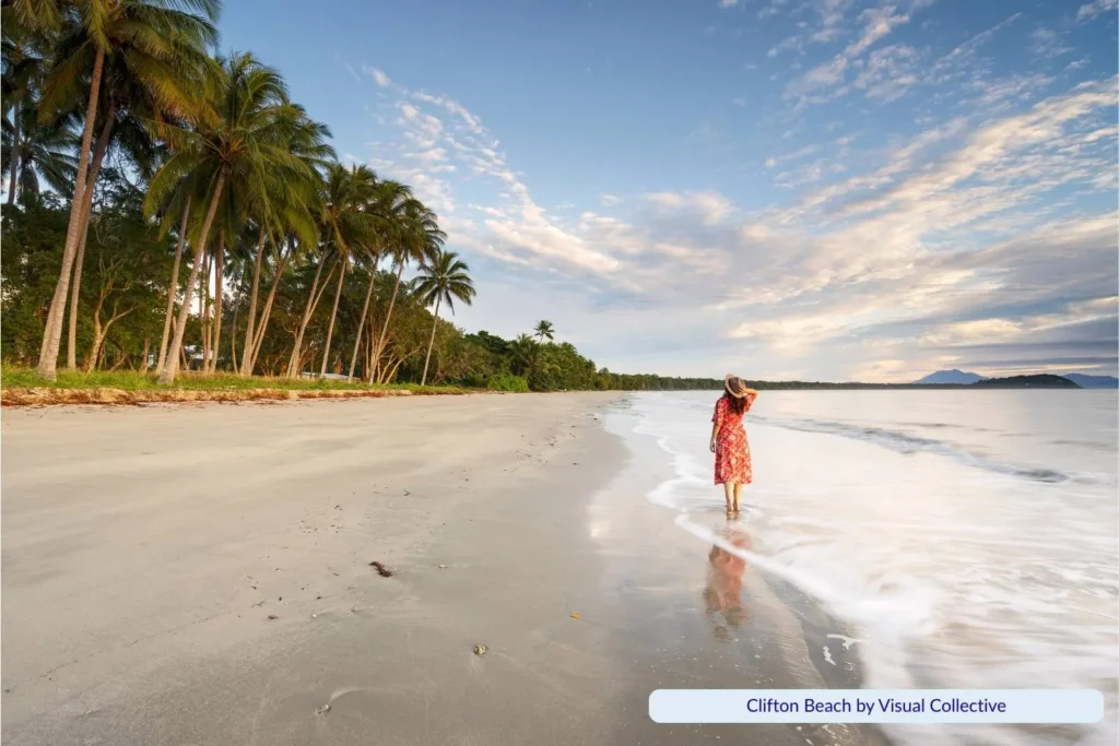 Woman walking along the shoreline at Clifton Beach in Cairns, Queensland, with soft waves washing onto the wide sandy beach lined with tall palm trees at sunset.