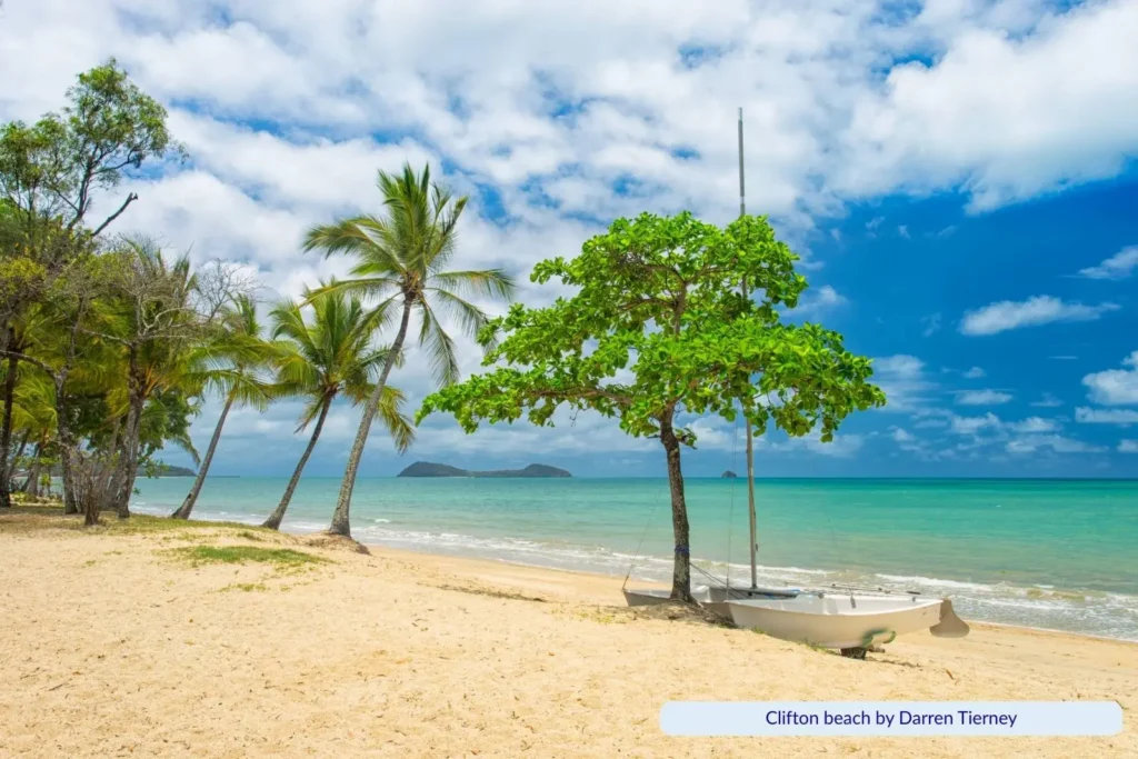 Clifton Beach in Cairns, Queensland, with golden sand, palm trees, and a small sailboat resting near the shoreline against turquoise waters and a partly cloudy sky.