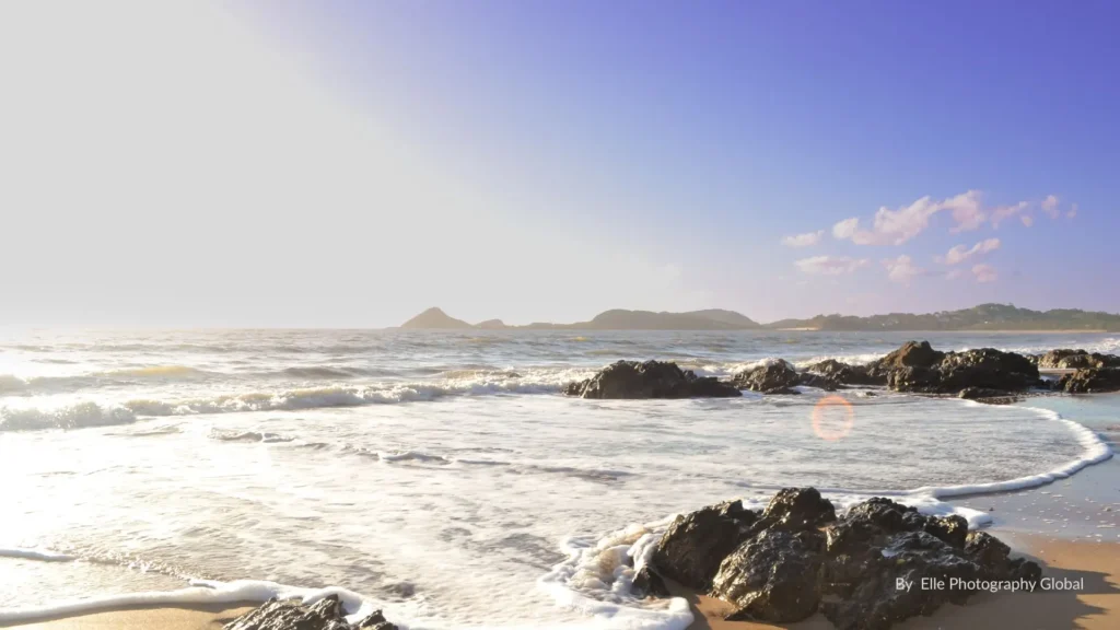 Rocky shoreline at Cooee Bay near Yeppoon, Queensland, with gentle waves and coastal hills in the distance under a bright sky.