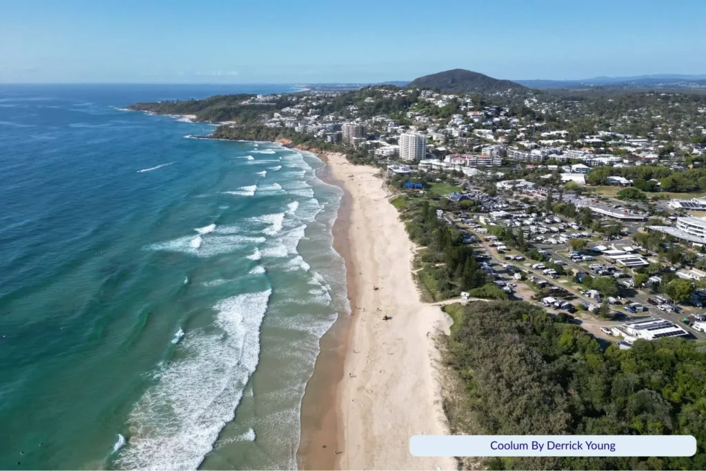 Aerial view of Coolum Beach on the Sunshine Coast, Queensland, showing golden sand, rolling surf waves, and the coastal township with green headland and hills in the background.