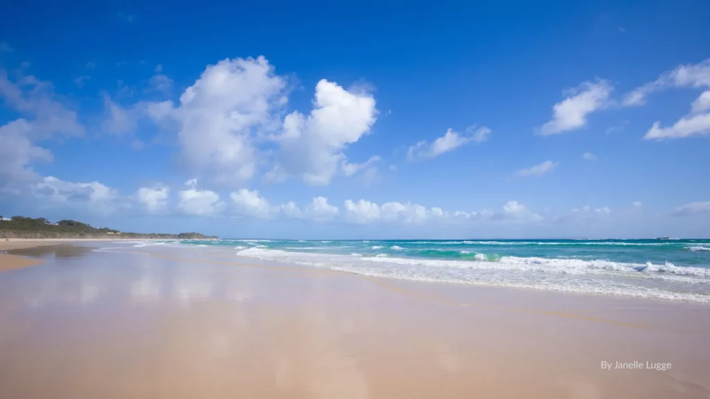 Wide sandy shoreline at Home Beach with turquoise waves, clear skies, and gentle surf, North Stradbroke Island, Queensland.