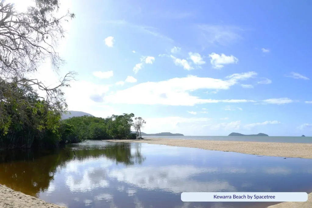 Scenic view of Kewarra Beach in Cairns, Queensland, with calm water lagoon reflecting the sky, sandy shoreline, and lush green trees leading out to the Coral Sea.