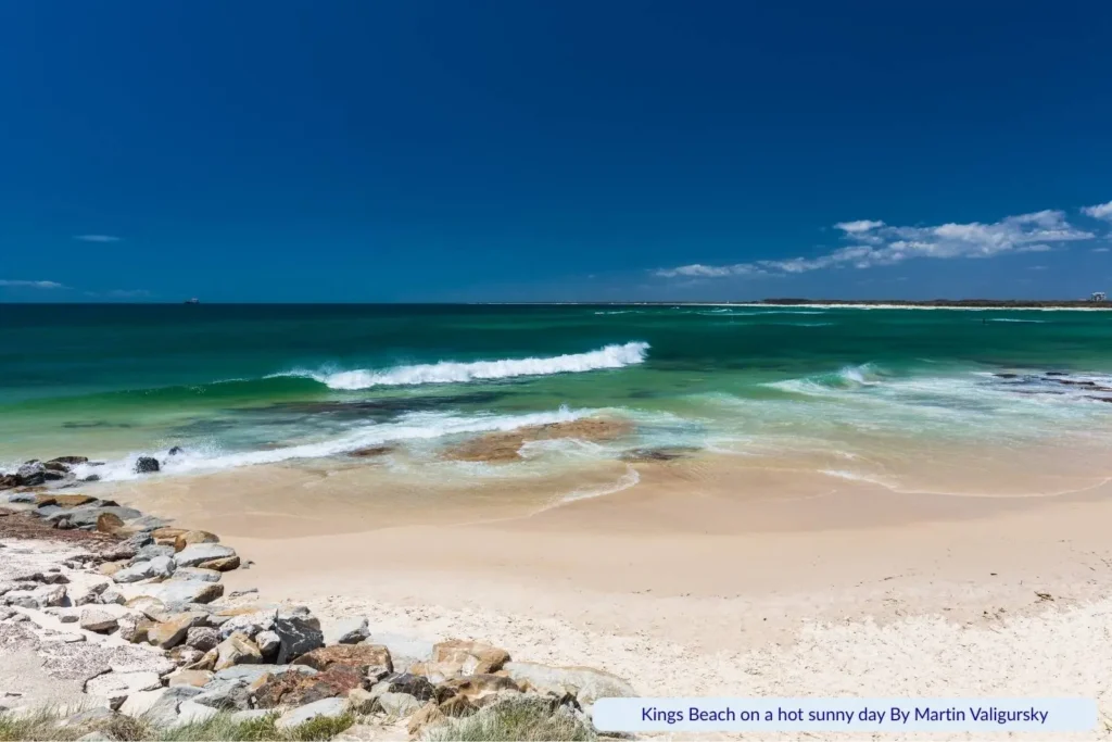 Kings Beach in Caloundra, Queensland, on a hot sunny day, with turquoise waves rolling onto golden sand and rocky edges under a clear blue sky.