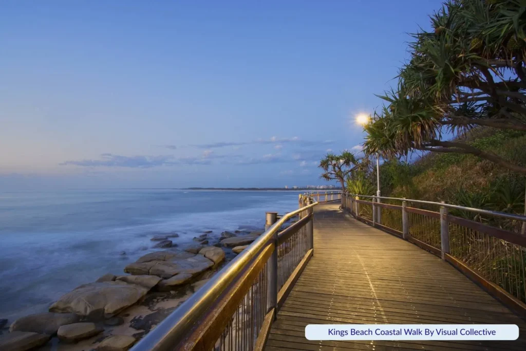 Kings Beach coastal boardwalk in Caloundra, Queensland, at dusk, with pandanus trees, rocky foreshore, and ocean views under a soft blue sky