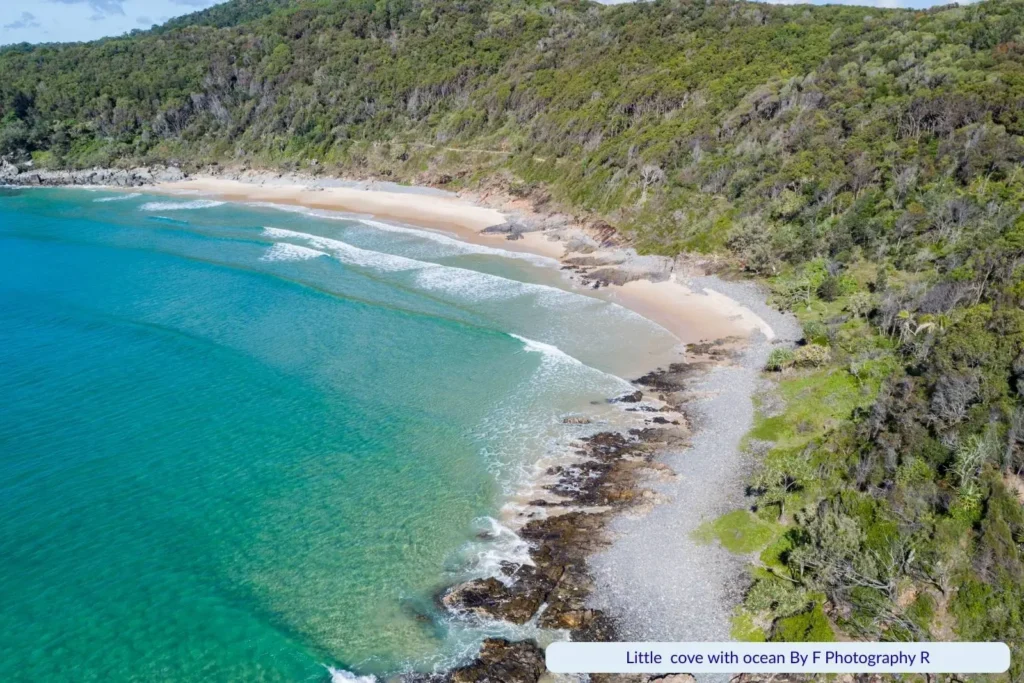 Aerial view of Little Cove Beach in Noosa, Queensland, with turquoise water, small waves rolling onto a sandy cove, rocky edges, and lush green forest hillside.