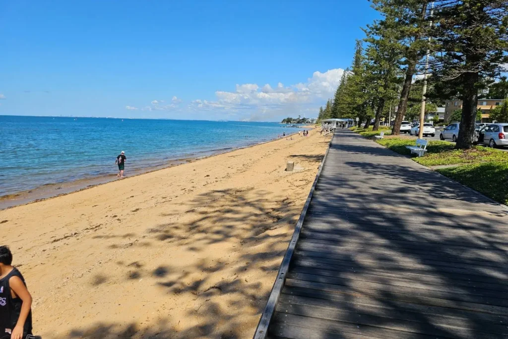 View along the timber boardwalk at Margate Beach with pine trees, beachgoers, and bright sunshine along the Brisbane coastline.
