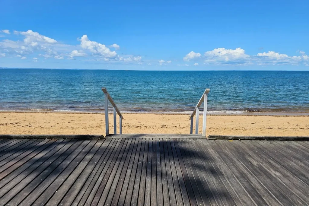 Wooden boardwalk leading to the golden sands and calm waters of Margate Beach, with a clear view of Moreton Bay under a bright blue sky in Brisbane, Queensland.