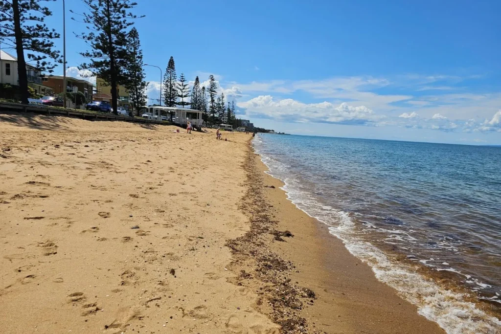 Paved walkway beside sandy Redcliffe Beach with calm blue water and pine trees lining the foreshore, Brisbane, Queensland.