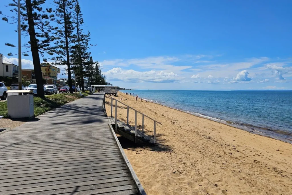 Elevated timber boardwalk beside golden sands and calm waters at Margate Beach, lined with tall pine trees and coastal buildings, Brisbane, Queensland.