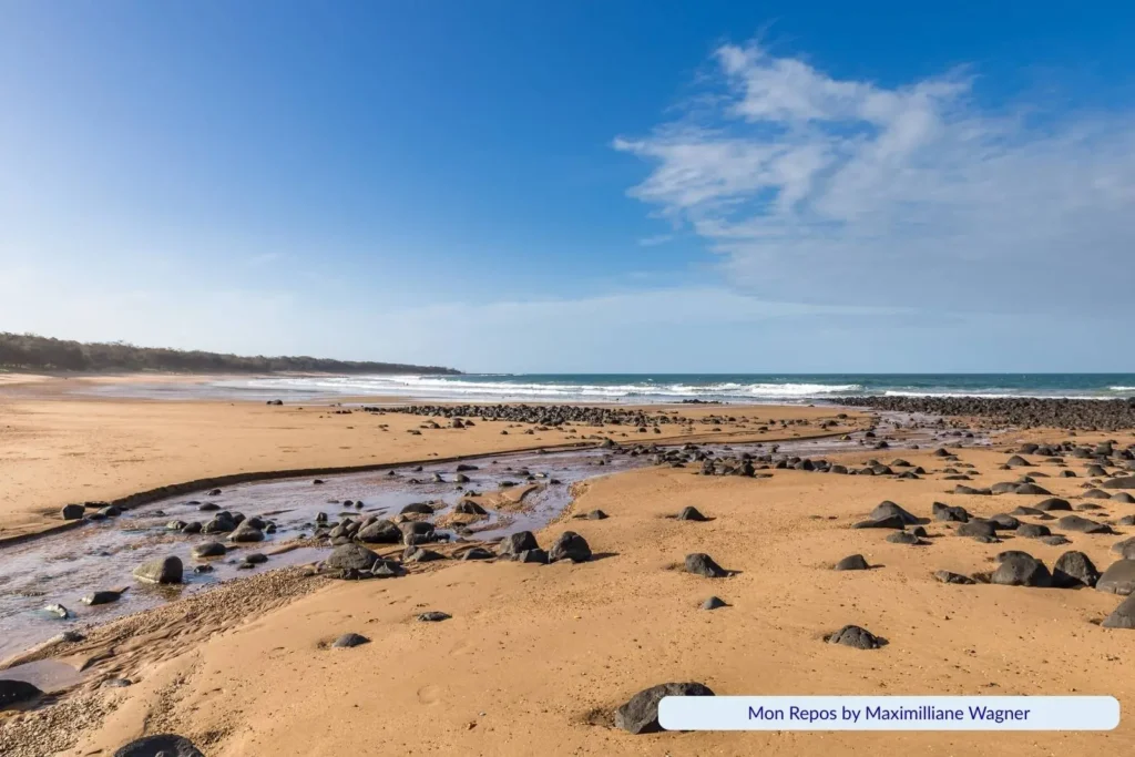 Wide view of Mon Repos Beach in Bundaberg, Queensland, with golden sand scattered with volcanic rocks, a small stream flowing to the ocean, and waves under a partly cloudy sky.