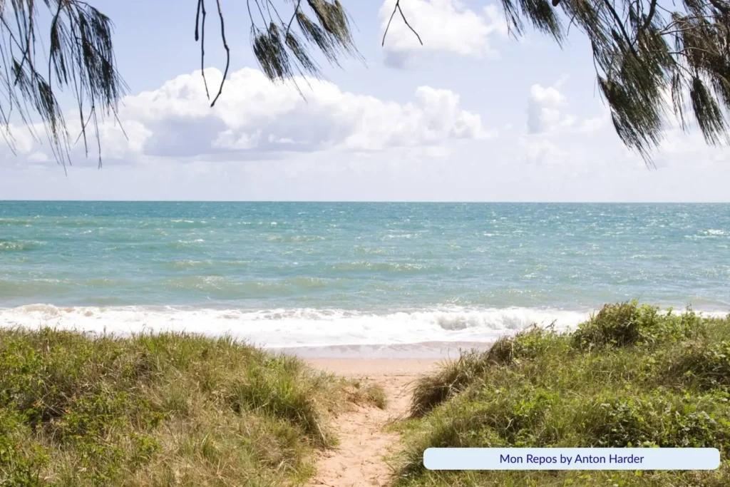 Pathway through grassy dunes leading to Mon Repos Beach in Bundaberg, Queensland, with turquoise ocean waves and cloudy blue sky framed by overhanging branches