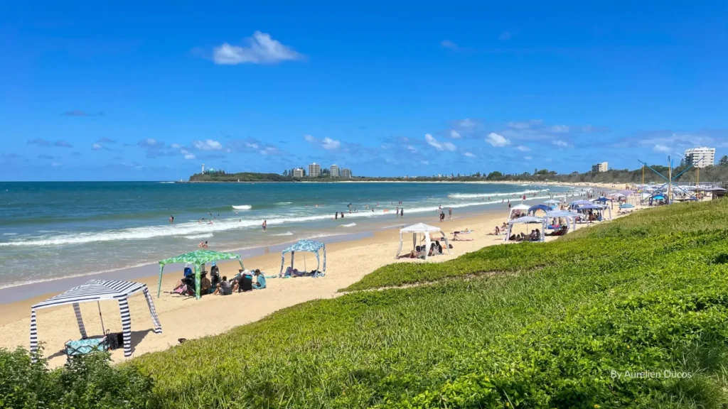 Mooloolaba Beach with golden sand, sunshades, swimmers in calm blue water, and high-rise apartments along the foreshore, Sunshine Coast, Queensland.