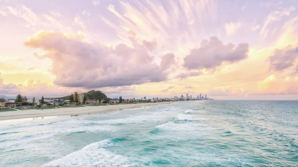Sunset over Nobby Beach with golden sand, gentle surf, and a pastel sky, with the Gold Coast skyline visible in the distance, Queensland, Australia.