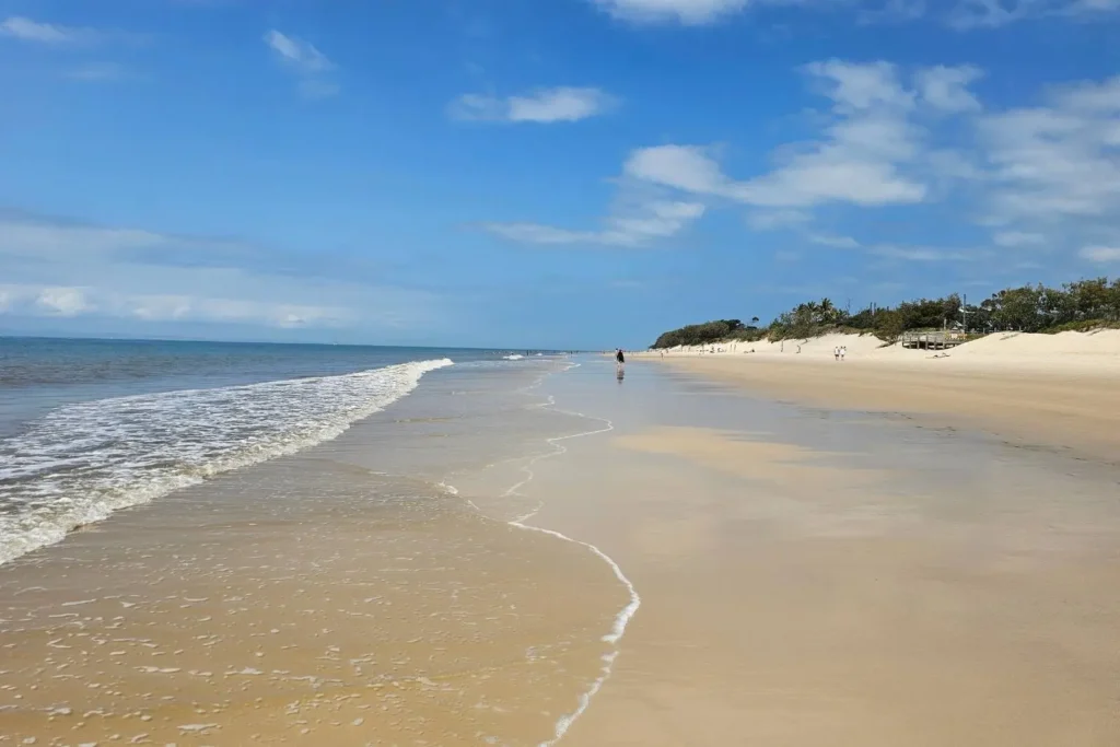 Shallow waves washing onto the wide sandy shoreline of Ocean Beach with people walking in the distance under a bright blue sky, Bribie Island, Queensland.