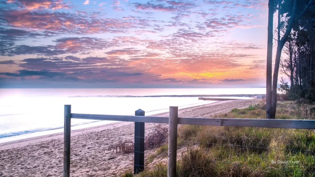 Sunset over Pialba Beach in Hervey Bay, with sandy foreshore, grassy parklands, and coastal trees in the foreground, Queensland.