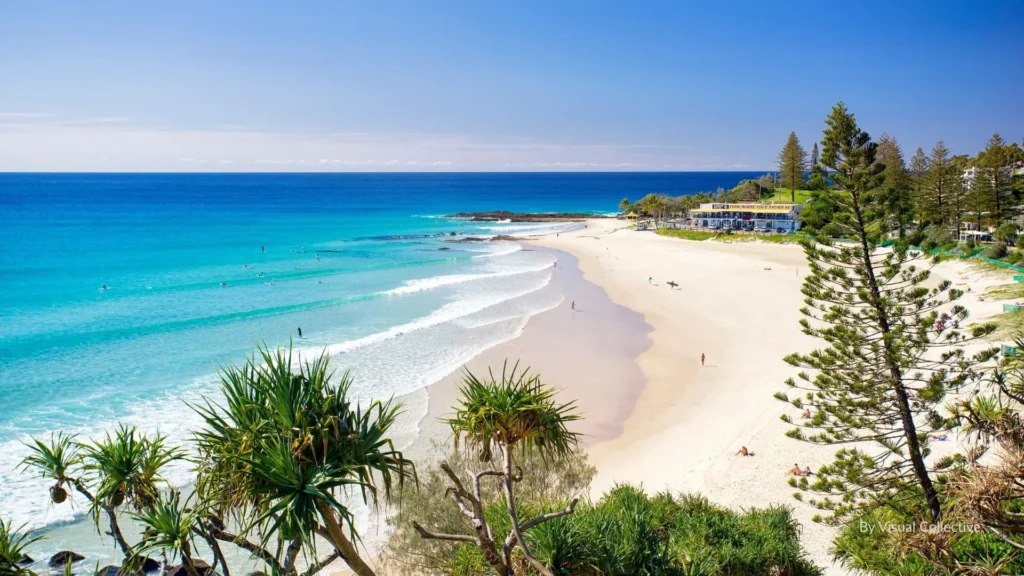Rainbow Bay with calm turquoise waters, golden sandy beach, and pandanus trees in the foreground, with Snapper Rocks visible in the distance, Gold Coast, Queensland, Australia.