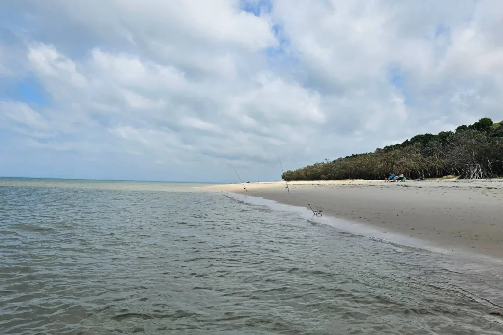 Shallow tidal waters at Red Beach on Bribie Island QLD, with sandy shoreline, fishing rods along the beach, and coastal bushland under a cloudy sky.