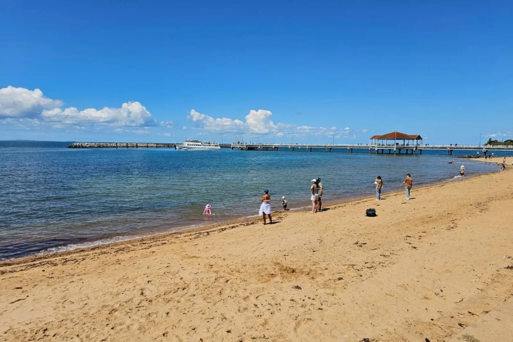 Redcliffe Beach with families enjoying the sandy shore and clear blue waters, with the historic Redcliffe Jetty in the background on a sunny day, Brisbane, Queensland.