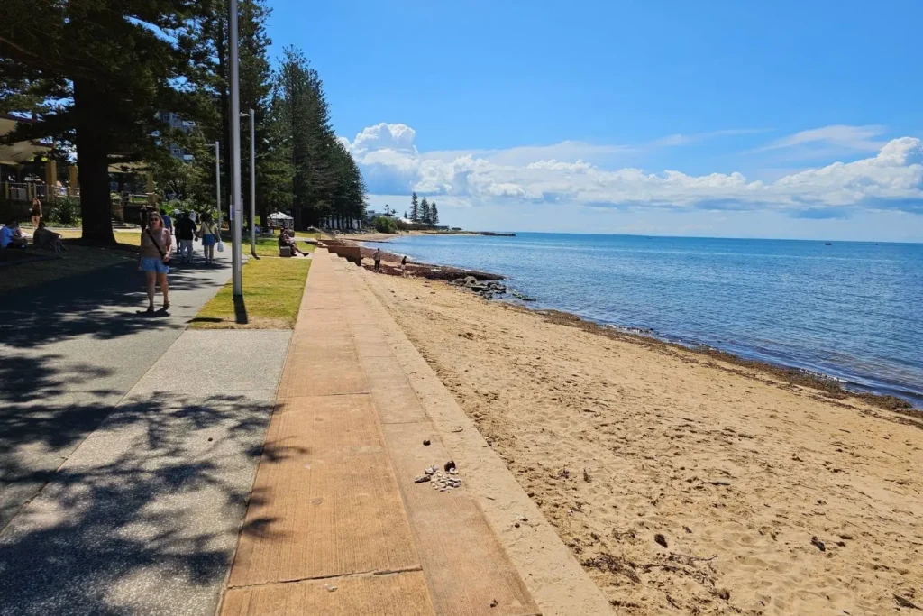 Scenic view of Redcliffe beach with calm waters, golden sand, and a paved coastal walkway lined with pine trees and people enjoying the sunshine, Moreton Bay Region, Queensland.