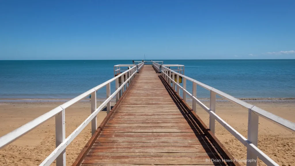 Wooden jetty stretching over golden sand into calm blue waters at Scarness Beach, Hervey Bay, Queensland.