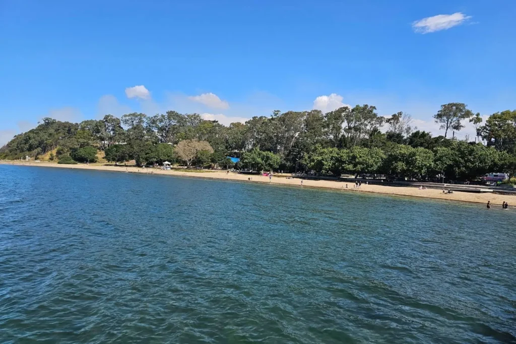 Shorncliffe Beach in Brisbane with calm blue water, tree-lined foreshore, and scattered visitors enjoying the quiet sandy shoreline under a bright blue sky.