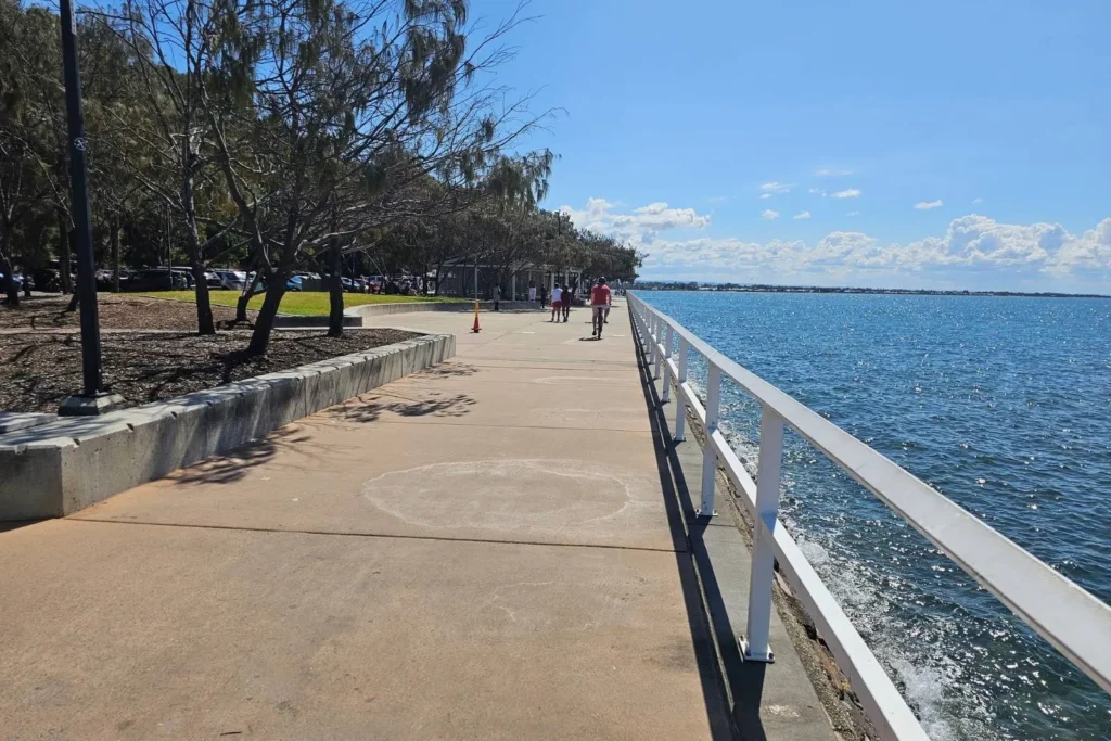 Scenic waterfront walkway at Shorncliffe, Brisbane, with railings along the calm bay, shaded trees to the left, and people strolling under a bright blue sky.