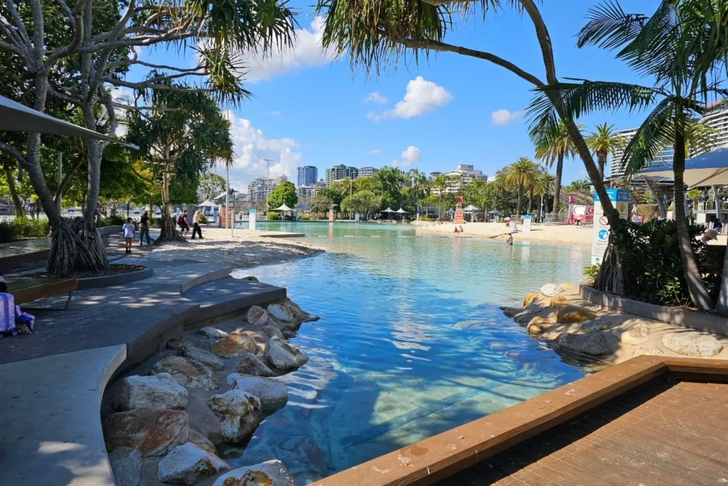 Streets Beach, South Bank Parklands, Brisbane QLD, Australia — man-made lagoon with palm trees, sandy shoreline and city skyline on a sunny day.
