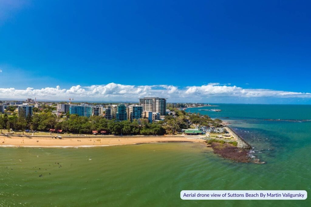 Aerial view of Suttons Beach in Redcliffe, Queensland, showing calm green waters, golden sand, high-rise buildings along the foreshore, and a clear blue sky stretching over Moreton Bay.