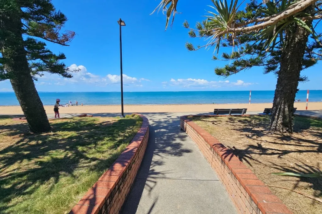 Accessible walkway leading to golden sand and calm blue waters at Suttons Beach, Redcliffe Peninsula, with shaded park area and ocean horizon under a clear sky.