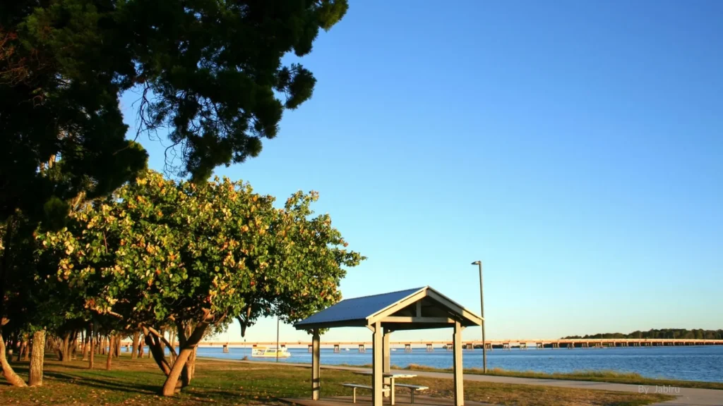 Scenic view of Sylvan Beach on Bribie Island, Queensland, with shaded picnic shelter, lush trees, and calm blue waters overlooking the Pumicestone Passage and Bribie Island Bridge.