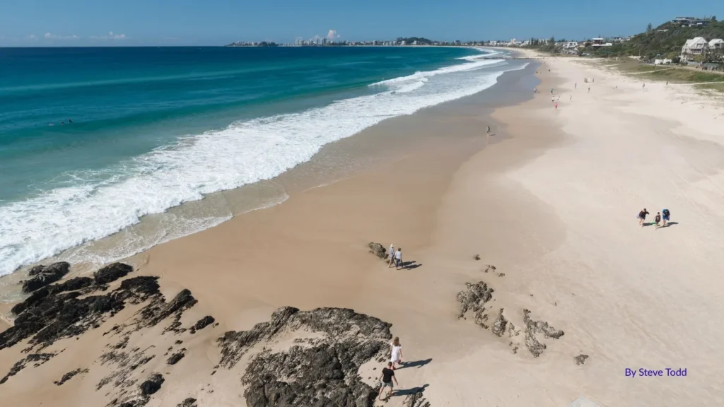 Wide sandy stretch of Tugun Beach with turquoise waves, rocky headland foreground, and people enjoying the uncrowded shoreline, Gold Coast, Queensland.