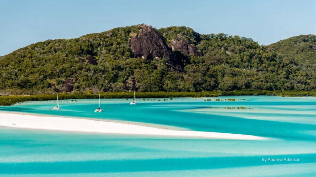 Turquoise waters and white silica sand with yachts anchored at Whitsunday Island, backed by lush green rainforest and rocky headlands, Queensland, Australia.