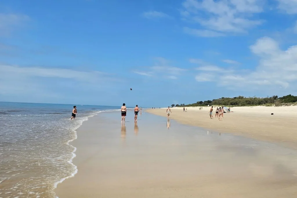 Visitors enjoying Woorim Beach on Bribie Island QLD, with people walking and playing along the wet sandy foreshore under a bright blue sky.