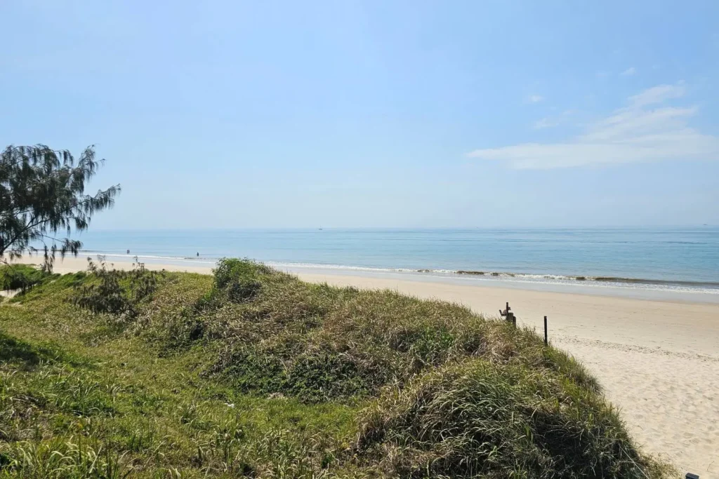 Golden sandy shoreline at Woorim Beach on Bribie Island QLD, with grassy dunes in the foreground, calm blue ocean, and people walking along the water’s edge