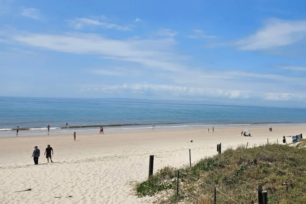 Visitors enjoying Woorim Beach on Bribie Island QLD, with golden sand, calm blue waves, grassy dunes, and families walking and relaxing along the shoreline.