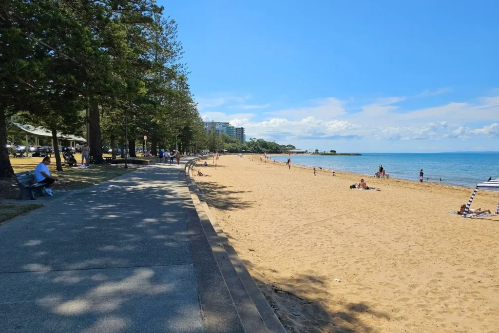 Shaded footpath alongside the golden sands of Suttons Beach, with beachgoers enjoying calm waters under a clear blue sky in Redcliffe, Queensland.