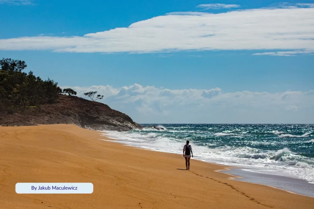 Person walking along the golden sands of Agnes Water Beach with gentle surf and blue skies, Southern Great Barrier Reef coastline, Queensland.