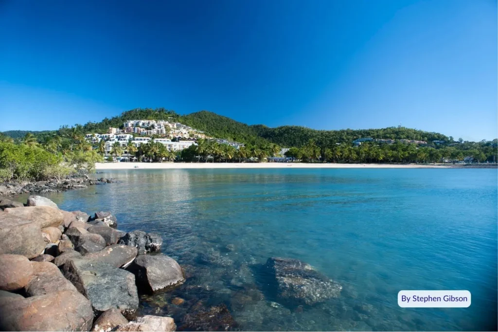 Rocky shoreline and calm blue bay with hillside resorts at Airlie Beach, gateway to the Whitsunday Islands, Queensland