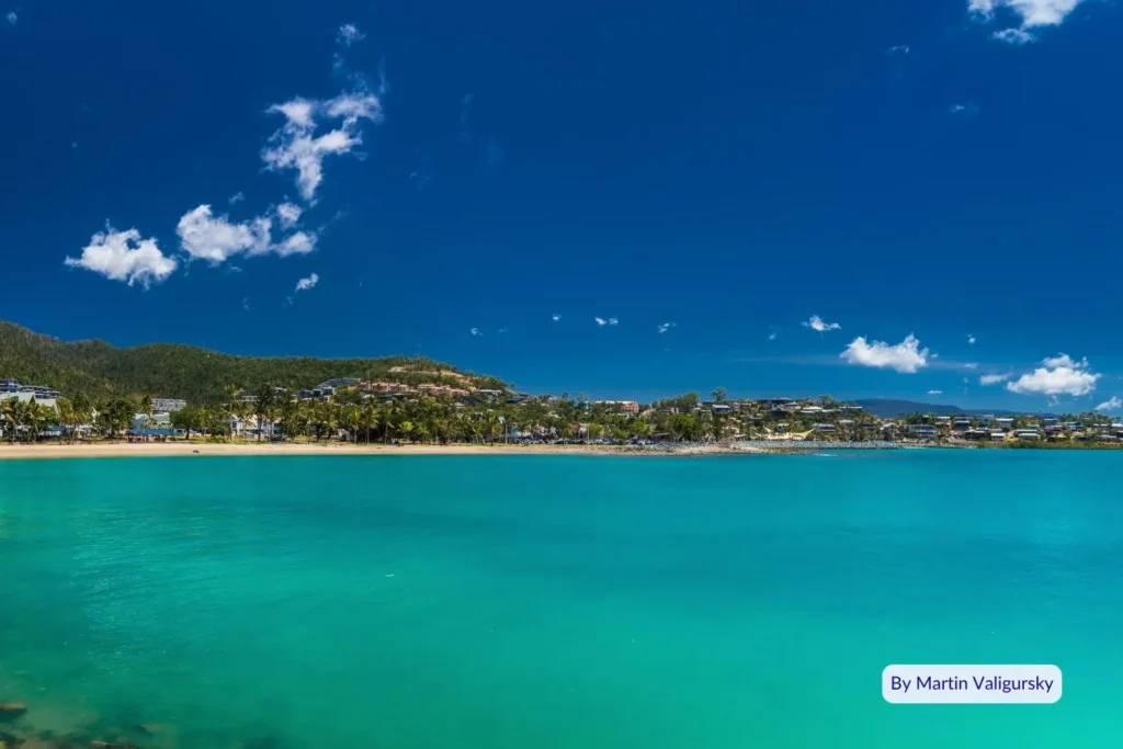 Panoramic view of Airlie Beach waterfront with clear turquoise water and tropical hillside town under bright blue sky, Queensland.