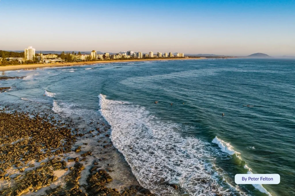 Aerial view of Alexandra Headland Beach showing surfers in turquoise waves, golden sand, and the Sunshine Coast skyline at sunrise, Queensland.