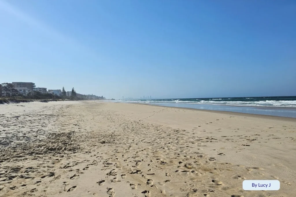 Golden sand stretching along Bilinga Beach, Gold Coast, with distant city skyline, footprints in the sand, and soft morning light.